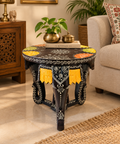 A round black accent side Elephant Head Table with intricate white hand-painted patterns, yellow tassels, and colorful floral motifs on the top, styled in a warm living room.