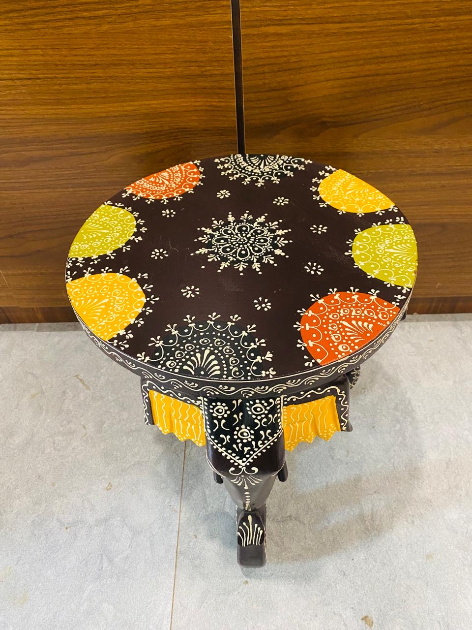 A round black accent side Elephant Head Table with intricate white hand-painted patterns, yellow tassels, and colorful floral motifs on the top, styled in a warm living room.